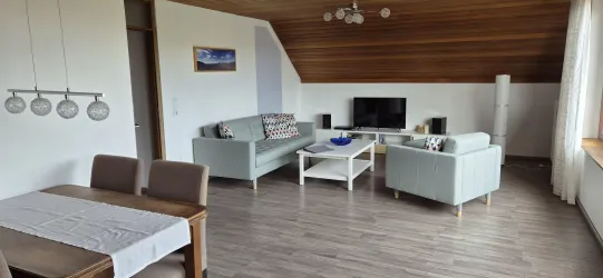 Ein modernes Wohnzimmer mit hellen Möbeln und einem großen Fernseher. Es gibt einen Essbereich mit einem Tisch und Stühlen.