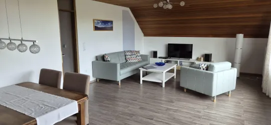 Ein modernes Wohnzimmer mit hellen Möbeln und Holzdecke. Es gibt eine gemütliche Sitzecke und einen Essbereich mit einem Tisch.