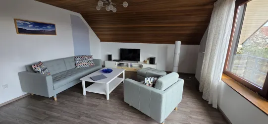 Ein modernes Wohnzimmer mit einem hellgrünem Sofa und einem bequemen Sessel. Es gibt einen Fernseher, einen Couchtisch und viel Tageslicht, das durch das Fenster strömt.