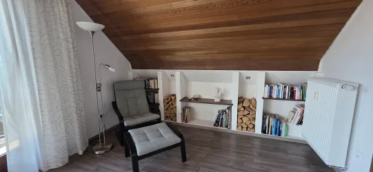 Ein gemütlicher Raum mit schrägem Holzdach. Ein bequemer Stuhl und ein Bücherregal sind zu sehen.