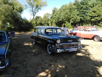 Ein klassischer schwarzer Kombi steht auf einem sonnigen Feld. Im Hintergrund sind weitere Oldtimer und Bäume zu sehen.
