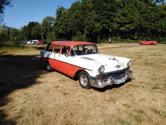 Ein klassischer, rot-weißer Kombi steht auf einer grünen Wiese. Im Hintergrund sind Bäume und weitere Fahrzeuge zu sehen.