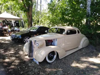 Ein klassisches Auto in Beige steht unter Bäumen. Im Hintergrund ist ein schwarzes Fahrzeug und ein Verkaufsstand zu sehen.