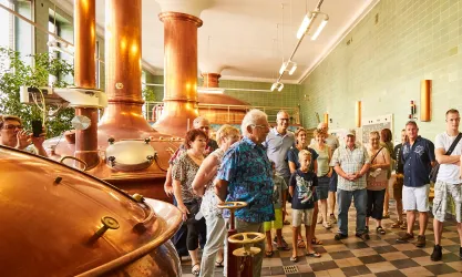 Eine Gruppe von Menschen steht in einer Brauerei und hört einer Führung zu. Im Hintergrund sind große kupferne Braukessel zu sehen.