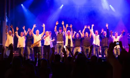 Eine Gruppe von Musikern steht auf einer Bühne und hebt die Hände in die Luft. Die Atmosphäre ist festlich mit bunten Lichtern im Hintergrund.
