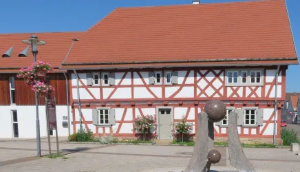 Ein altes Fachwerkhaus mit rotem Dach und weißen Wänden steht in einem ruhigen Platz. Neben dem Haus befindet sich eine moderne Skulptur und blühende Pflanzen.