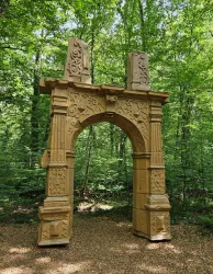 Ein kunstvoll gestalteter Torbogen aus Holz steht im Wald. Die umgebende Natur ist grün und einladend.