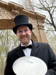 Ein Mann in einem Zylinder und einem Frack lächelt, während er ein Musikinstrument in der Hand hält. Im Hintergrund sind Bäume und eine dekorative Struktur zu sehen.