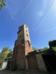 Ein alter, höherer Turm aus Ziegeln steht in einer ländlichen Umgebung. Der Himmel ist blau und klar, umgeben von Bäumen.