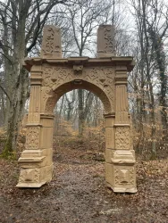 Ein dekorativer Bogen aus Stein steht in einem Wald. Umgeben von Bäumen und herbstlichem Laub.