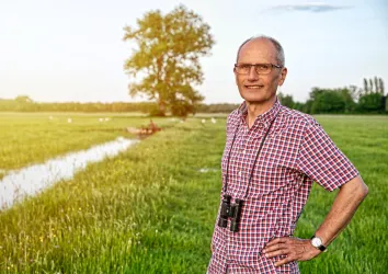 Ein Mann steht auf einer Wiese mit einem Fernglas um den Hals. Im Hintergrund ist ein Baum und ein Wasserlauf zu sehen.