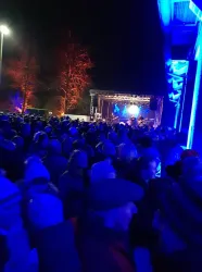 Ein großes Publikum versammelt sich vor einer Bühne bei Nacht. Die Atmosphäre ist festlich mit bunten Lichtern und Musik.