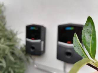 Zwei schwarze Geräte an einer Wand, möglicherweise Sensoren oder Steuerungen für eine smarte Anwendung. Im Vordergrund ist ein grünes Blatt mit Wassertropfen zu sehen.