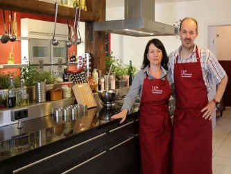 Ein modernes Küchenatelier mit zwei Personen in roten Schürzen. Die Küche ist gut ausgestattet, mit Pflanzen und Kochutensilien im Hintergrund.