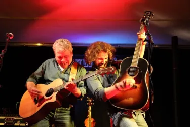 Zwei Musiker, die auf der Bühne spielen und Spaß haben. Beide spielen akustische Gitarren vor einer beleuchteten Kulisse.