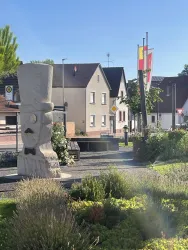 Eine Statue mit einem Hut und einem Schnurrbart steht in einem kleinen Platz. Im Hintergrund sind Häuser und Fahnen sichtbar.