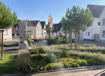 Eine ruhige Parklandschaft mit einem modernen Kunstwerk und kleinen Bäumen. Im Hintergrund sind Wohnhäuser und Flaggen sichtbar.