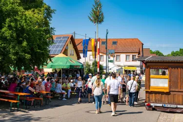 Eine belebte Straße bei einer Veranstaltung mit vielen Menschen. Es gibt bunte Zelte und Tische, die zum Verweilen einladen.