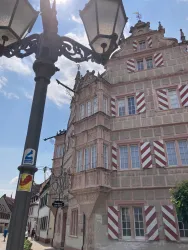 Ein historisches Gebäude mit einer auffälligen Fassade in sanften Farben. Im Vordergrund ist eine Straßenlaterne sichtbar.