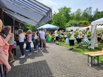 Eine Menschenmenge bei einer Veranstaltung im Freien. Im Hintergrund steht eine Musikkapelle in bunten Kostümen.