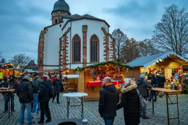 Ein Weihnachtsmarkt mit festlich dekorierten Ständen und vielen Besuchern. Im Hintergrund ist eine Kirche zu sehen, die die winterliche Atmosphäre unterstreicht.