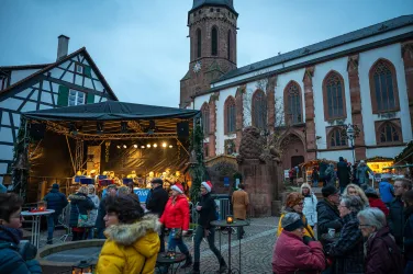 Ein lebhafter Weihnachtsmarkt mit vielen Menschen und festlicher Atmosphäre. Im Hintergrund steht eine Kirche und eine Bühne mit Musik.