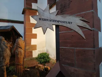 Ein dekoratives Schild mit der Aufschrift "Weihnachtspostamt" in Form eines Sterns. Im Hintergrund sind geschnitzte Figuren und etwas Grün zu sehen.