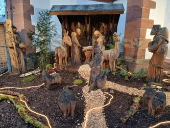 Eine Schnitzerei mit Figuren aus Holz, die eine Nativity-Szene darstellen. Umgeben von Pflanzen und einer kleinen Lichterkette.