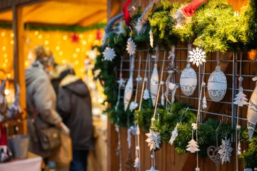 Ein festlicher Weihnachtsmarkt mit geschmückten Wänden aus Tannenzweigen und ornamentalen Dekorationen. Im Hintergrund sind unscharfe Personen zu sehen, die den Markt erkunden.
