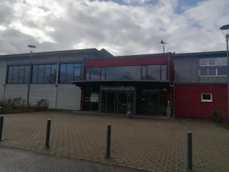 Ein modernes Gebäude mit einer großen Glasfront und einem Eingang. Die Fassade ist teilweise rot und die Umgebung hat einen gepflasterten Boden.