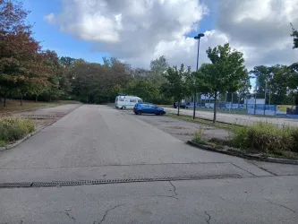 Ein leerer Parkplatz mit vereinzelten Autos und Bäumen im Hintergrund. Die Wolken sind teilweise bedeckt und es ist eine ruhige Atmosphäre zu sehen.
