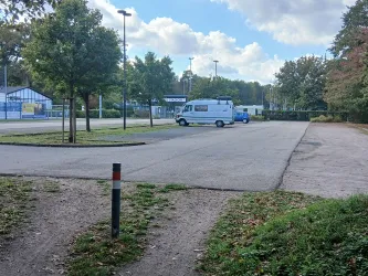 Ein leeres Parkplatzgelände mit wenigen Fahrzeugen. Im Hintergrund sind einige Bäume und Gebäude sichtbar.