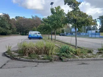 Eine ruhige Parkfläche mit vereinzelten Fahrzeugen und einigen Bäumen. Im Hintergrund sind Sporteinrichtungen sichtbar.