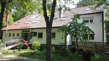 Ein modernes Gebäude umgeben von Bäumen und blühenden Sträuchern. Es hat ein rotes Satteldach und eine Zugangsrampe.