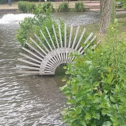 Eine künstlerische Holzstruktur in Form eines Fächers steht im Wasser. Umgeben von grünem Blattwerk strahlt die Szene Ruhe und Naturverbundenheit aus.