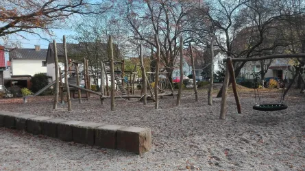 Ein Spielplatz mit Holzspielgeräten und einer Schaukel. Der Sandboden ist von Bäumen umgeben.