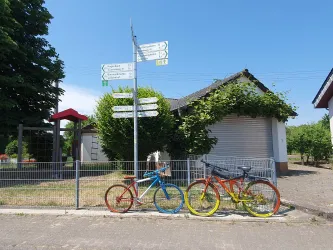 Zwei farbige Fahrräder stehen an einem Zaun vor einem Gebäude. Im Hintergrund sind Wegweiser und eine grüne Hecke zu sehen.