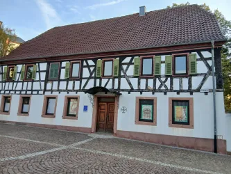 Ein historisches Fachwerkhaus mit einem roten Ziegeldach und grünen Fensterläden. Der Eingangsbereich ist schön gestaltet und die Fassade hat einen klaren weißen Farbton.