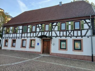 Ein traditionelles Fachwerkhaus mit grünem Fensterladen. Die Fassade ist weiß mit einem Ziegeldach und liegt an einem gepflasterten Platz.