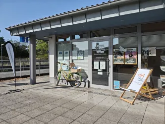 Ein modernes Informationszentrum mit großen Fenstern und einem Fahrrad vor der Tür. Auf dem Bürgersteig steht ein aufklappbarer Gästeständer mit Informationen.
