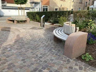 Ein moderner Platz mit geschwungenen Bänken aus Holz und Stein. Umgeben von sorgfältig gepflegten Pflanzen und Büschen.