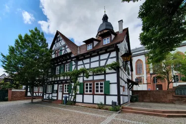 Ein gemütliches Fachwerkhaus mit einem hohen Dach und grünem Fensterladen. Im Hintergrund sind Bäume und ein Teil einer Kirche sichtbar.