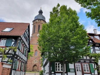 Eine malerische alte Stadt mit Fachwerkhäusern und einer hohen Kirche im Hintergrund. Im Vordergrund steht ein großer Baum, der Schatten spendet.