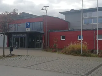 Ein modernes Gebäude mit roter Fassadenfarbe und großen Fenstern. Es gibt einen Parkplatz und einige Grünflächen in der Nähe.