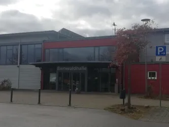 Die Bienwaldhalle ist ein modernes Gebäude mit großen Fenstern und roter Fassade. Im Vordergrund sind Parkplätze und einige Bäume zu sehen.