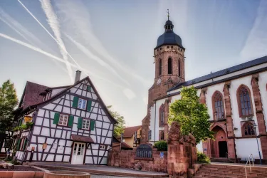 Eine malerische Altstadt mit einem Fachwerkhaus und einer imposanten Kirche. Der Himmel ist klar und es gibt sichtbare Flugstreifen.