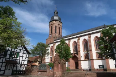 Eine Kirche mit einem markanten Turm und großen Fenstern. Im Vordergrund ist ein traditionelles Fachwerkhaus zu sehen, umgeben von Bäumen.
