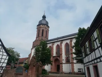 Eine historische Kirche mit einem hohen Turm steht neben traditionellen Fachwerkhäusern. Der Himmel ist bewölkt und die Umgebung ist grün.