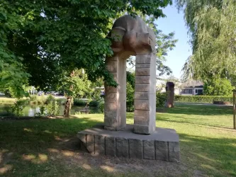Eine bemerkenswerte Steinskulptur steht unter schattigen Bäumen in einem Park. Im Hintergrund sieht man eine ruhige Grünfläche mit weiteren Kunstwerken.