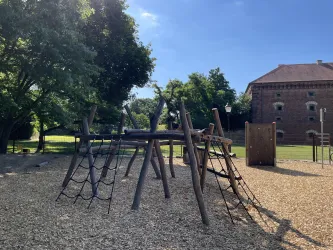 Ein Spielplatz mit einer Kletterstruktur aus Holz und einem weichen Boden aus Holzschnitzeln. Im Hintergrund sind Bäume und ein historisches Gebäude zu sehen.